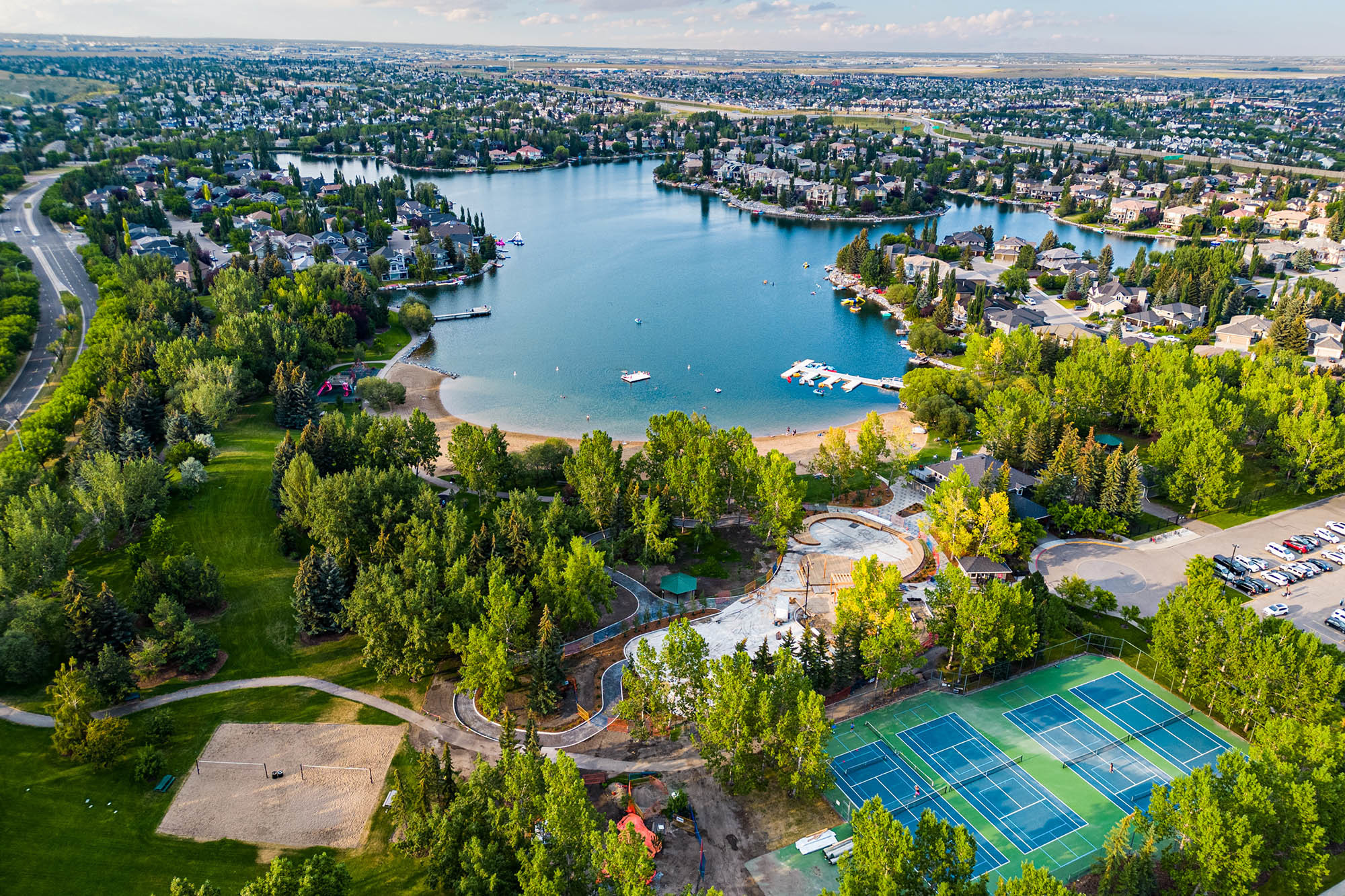 An aerial drone photo over McKenzie Lake in Southeast Calgary, a beautiful residential neighbourhood with tall trees, tennis courts, and lakefront homes for sale